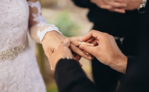 Två händer i svarta kostymärmar trär en vigselring på handen på en person i vit klänning. Foto: iStock
