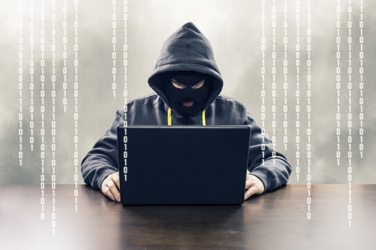 Bilden visar en person i luvtröja med dolt ansikte som sitter vid en bärbar dator. I bildbakgrunden anas "kod" som för tankarna till hackning.