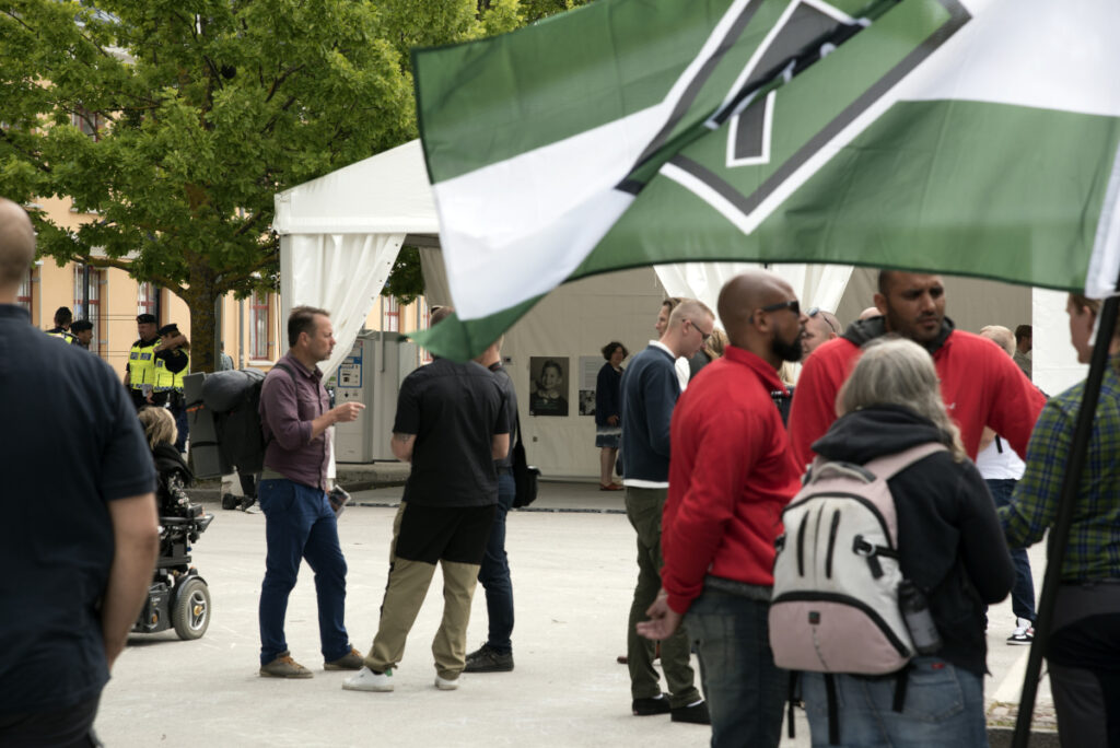 Bilden visar Nordiska Motståndsrörelsens tält i Almedalen, 2017. NMR:s flagga i förgrunden, Personer står runt omkring och diskuterar. En grupp poliser srtår i bakgrunden. Foto: Mattias Glassel, NU