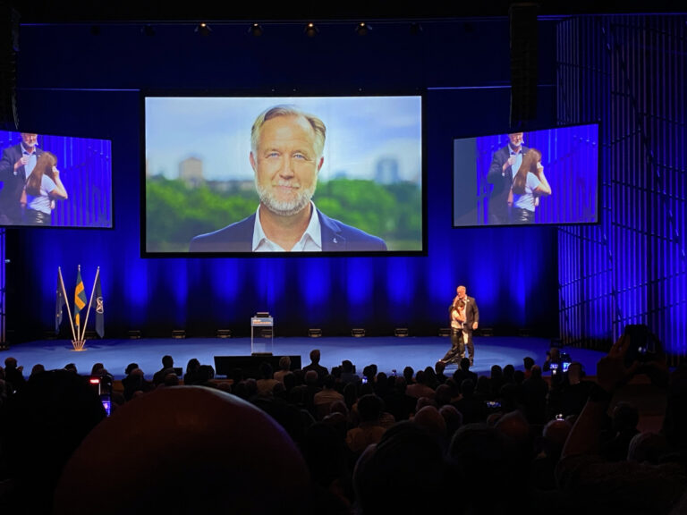 Johan Pehrson kommer upp på scenen för sitt tal, med flaggor i bakgrunden och sitt eget ansikte på storbild ovanför.