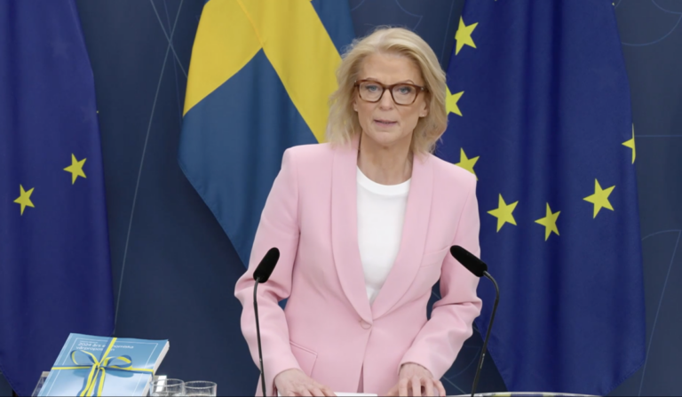 Elisabeth Svantesson i rosa kavaj presenterar vårbudgeten 2024, framför Sveriges och EU:s flaggor i bakgrunden.