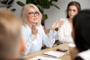 En kvinna i ljusblå skjorta leder en diskussion runt ett bord, omgivet av folk. Hon talar och slår ut med händerna. Foto: iStock