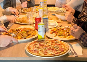 På bilden syns sju tallrika på ett bord, de flesta med olika pizzor på. Vi ser händerna på personer som äter pizza, brukar med läsk till, men inte personerna själva.