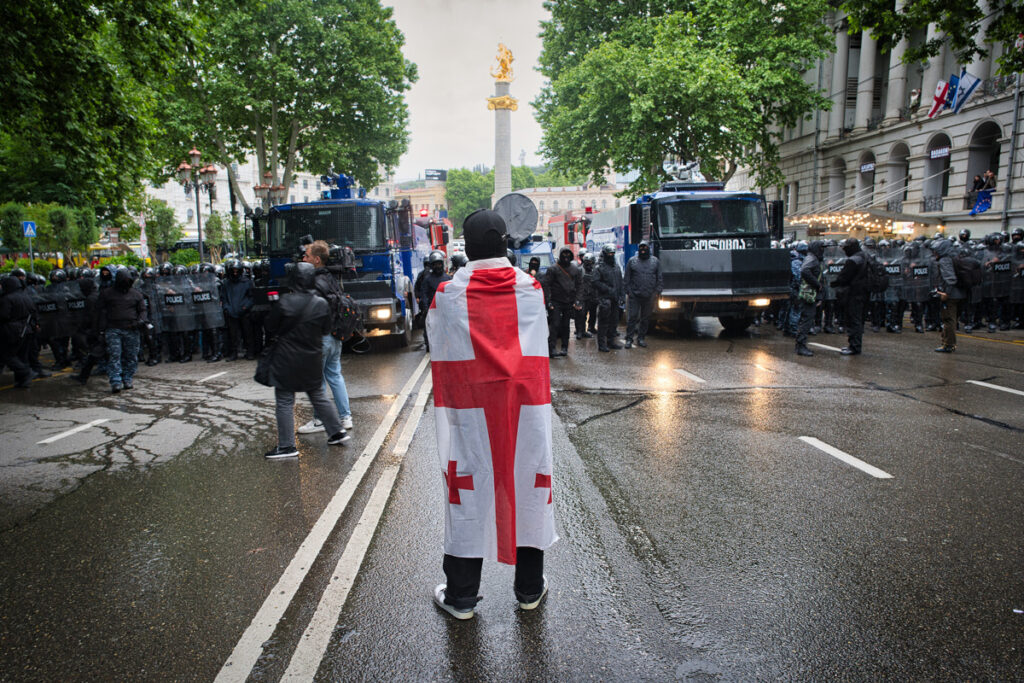 Bilden visar en man med Georgiens flagga drapperad över axlarna, som står mitt i vägen framför ett stort uppbåd kravallpoliser. Bilden tagen vid tidigare protester i Georgiens huvudstad. Foto: Rasmus Canbäck